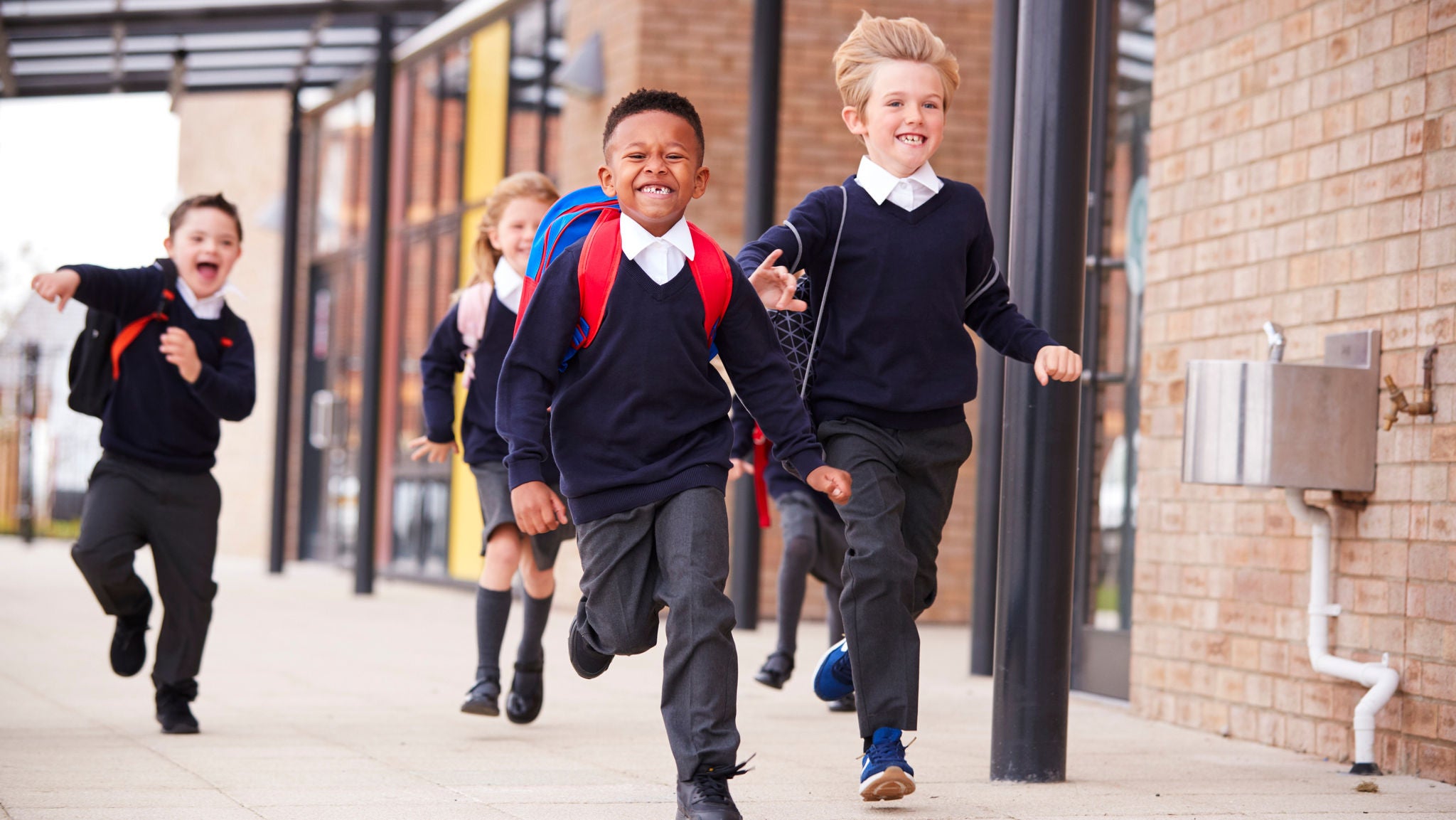 Happy primary school kids, wearing school uniforms and backpacks, running on a walkway outside their school building, front view, close up
